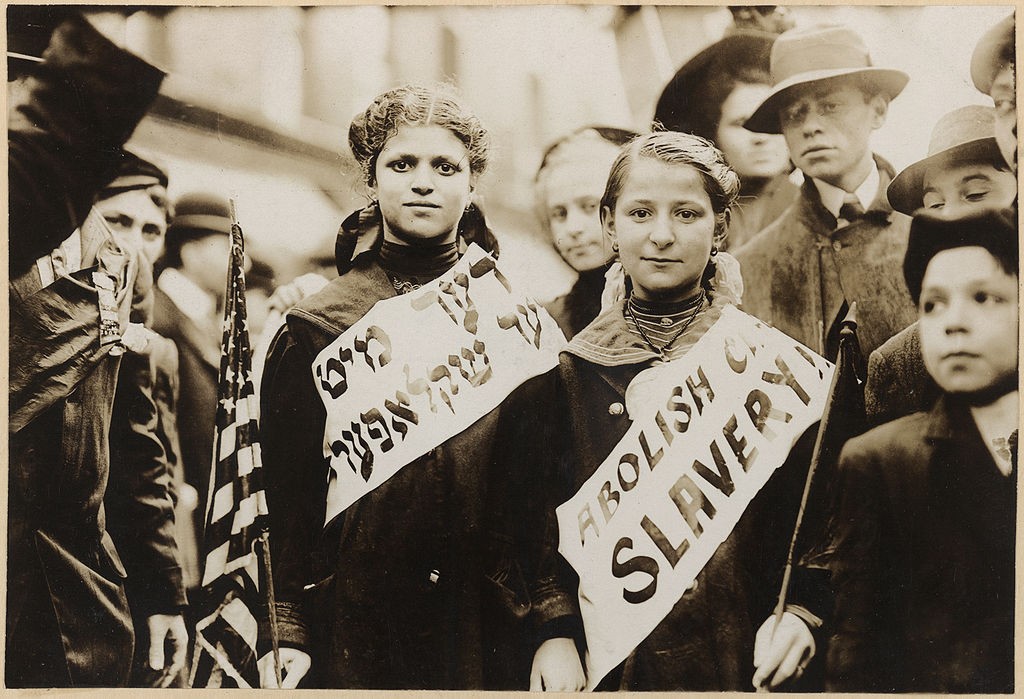 Photograph shows half-length portrait of two girls wearing banners with slogan "ABOLISH CH[ILD] SLAVERY!!" in English and Yiddish ("(ני)דער מיט (קינד)ער שקלאפער(ײ)", "Nider mit Kinder Schklawerii"), one carrying American flag; spectators stand nearby. Probably taken during May 1, 1909 labor parade in New York City.